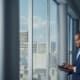 Thoughtful Black Businessman in a Tailored Suit Using Laptop while Standing in Office Near Window on Big City. Successful Corporate Top Manager Doing Data Analysis for e-Commerce Startup